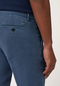 Blauwe linnen broek met een gestructureerde afwerking, voorzien van een achterzak en een knoopsluiting. Klein geborduurd hondenlogo boven de zak.