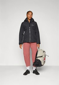 Zwarte waterdichte jas met een rits aan de voorkant, roze broek en een meerkleurige rugzak, staand tegen een effen achtergrond.