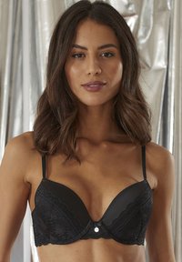 Reggiseno in pizzo nero con coppe imbottite, spalline regolabili, bordi scallop e un piccolo logo argento al centro.