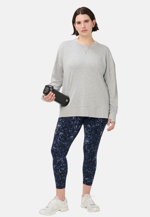 Vrouw staat en houdt een zwarte waterfles vast, draagt een lichtgrijze trui, donkerblauwe leggings met patroon en witte sneakers.