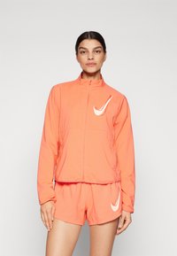Veste de sport orange avec fermeture éclair à l'avant et col haut, associée à un short assorti. Comprend un logo Nike blanc sur les deux pièces.