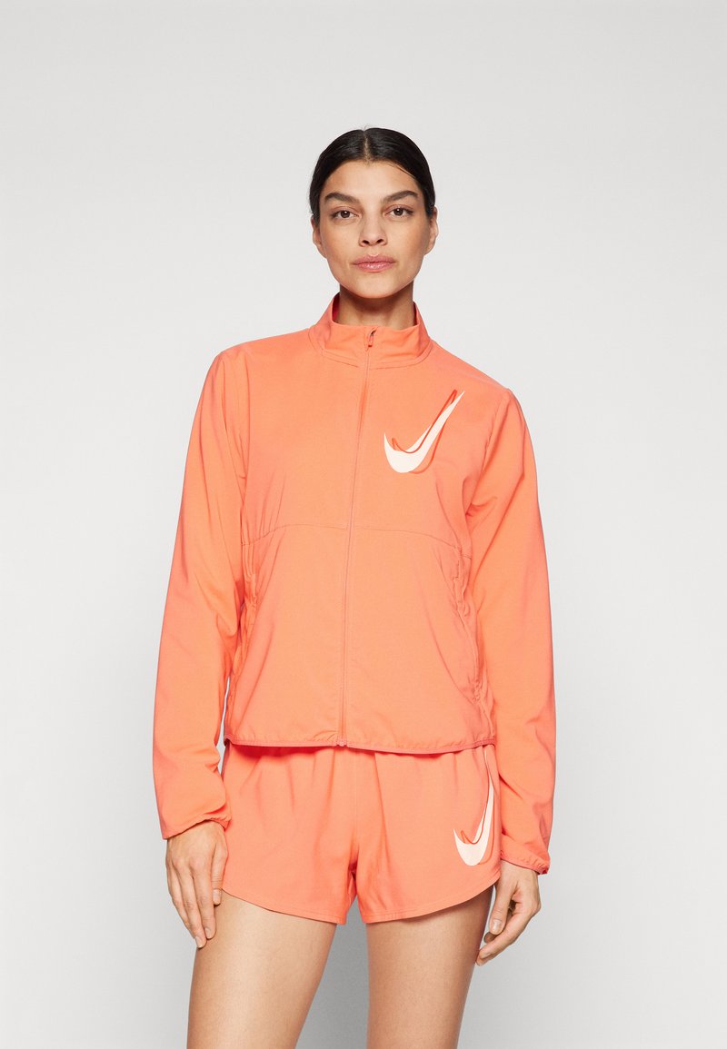 Veste de sport orange avec fermeture éclair à l'avant et col haut, associée à un short assorti. Comprend un logo Nike blanc sur les deux pièces.
