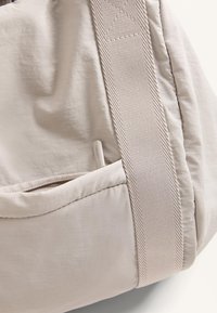 Beige tygväska med en mjuk, texturerad yta. Har ett horisontellt band och en synlig sidoficka med en liten etikett.