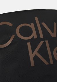 Svart texturerad väska med stora bruna bokstäver "Calvin Klein". Materialet verkar hållbart med en mjuk yta och framträdande branding.