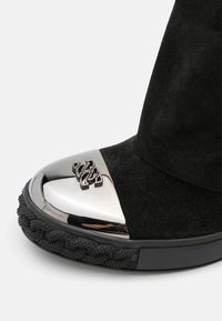 Botín de ante negro con puntera metálica brillante y emblema decorativo, suela negra texturizada y detalles trenzados.