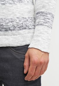 Grau-weiß gestreifter Strickpullover mit gerippten Bündchen, auf dem ein Hand auf dunklen Jeans ruht. Die Textur wirkt weich und warm.