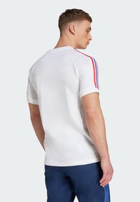 Uomo che indossa una camicia bianca a maniche corte con righe rosse e blu sulle maniche e pantaloni blu scuro con righe blu, girato di spalle.