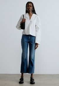 Fehér, rövid fazonú, V-nyakú ing, kék bővülő farmerek és fekete mokaszinek. Kiegészítők: textúrált fekete táska és karika fülbevalók.