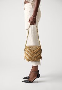 Persoon in off-white jeans en zwarte hakken, met een gestructureerde beige en gouden handtas met kettingstraps aan de zijkant.