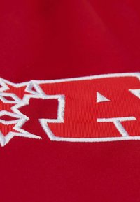 Roter Stoff mit weißer Stickerei. Das Design beinhaltet ein stilisiertes Logo mit geometrischen Formen und Akzenten, die Textur und Kontrast betonen.