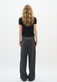 Top negro de manga corta combinado con pantalones grises sueltos y plisados. El cabello es largo y ondulado. Fondo blanco liso.