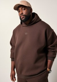 Uomo che indossa un felpa marrone con cappuccio, un cappello beige con logo e un orologio da polso, in piedi contro uno sfondo chiaro e semplice.