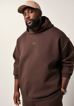 Man wearing a brown hoodie, beige cap with logo, and wristwatch, standing against a plain light background.