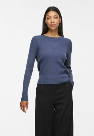 Femme aux longs cheveux foncés portant un pull côtelé bleu et un pantalon noir, debout, une main dans la poche, sur fond blanc.