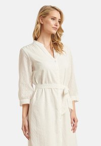 Abito in cotone bianco con chiusura a bottoni, maniche 3/4, cintura legata in vita e un motivo di ricami a occhio in tutto l'abito.