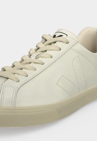 Gros plan d'une basket basse blanche avec des lacets beiges, une semelle en caoutchouc texturée et un logo V cousu sur le côté.