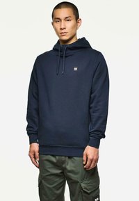 Uomo che indossa un hoodie blu navy con cordini e pantaloni cargo verde oliva, in piedi contro uno sfondo bianco semplice.