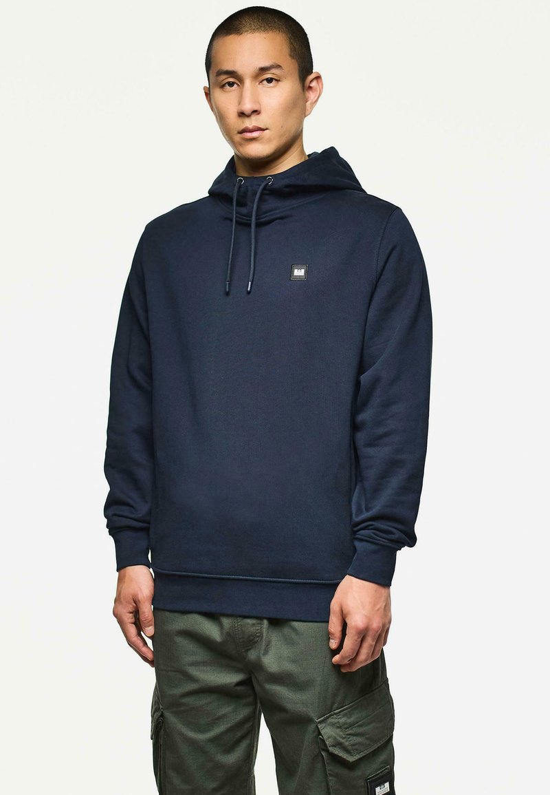 Uomo che indossa un hoodie blu navy con cordini e pantaloni cargo verde oliva, in piedi contro uno sfondo bianco semplice.
