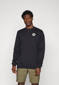 Sudadera negra con cuello redondo, con un parche de logo en el pecho. El material es suave, con puños y dobladillo acanalados. Combinada con pantalones cortos caqui.