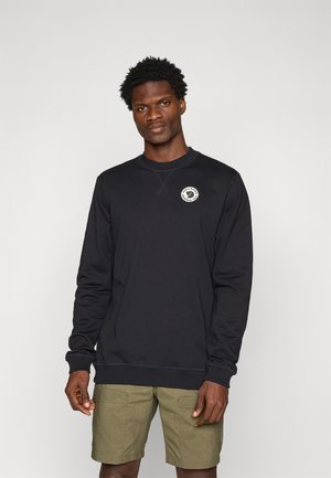 Schwarzer Sweater mit Rundhalsausschnitt, versehen mit einem Logo-Patch auf der Brust. Das Material ist glatt, mit gerippten Bündchen und Saum. Kombiniert mit khakifarbenen Shorts.