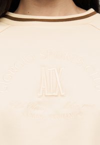 Μπεζ πουκάμισο από πίσω με κεντημένες λεπτομέρειες κειμένου «Giorgio Sports Club», «A|X», «Club House», «Milan 1991» και «Armani Exchange».