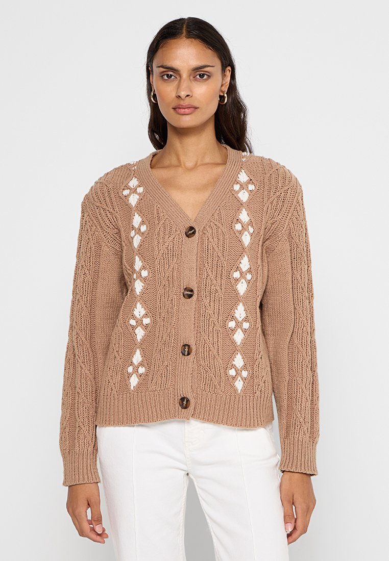 Beige gestrickte Strickjacke mit V-Ausschnitt, die Rautenausschnitte und strukturierte Muster aufweist, mit braunen Knöpfen verschlossen. Lange Ärmel.