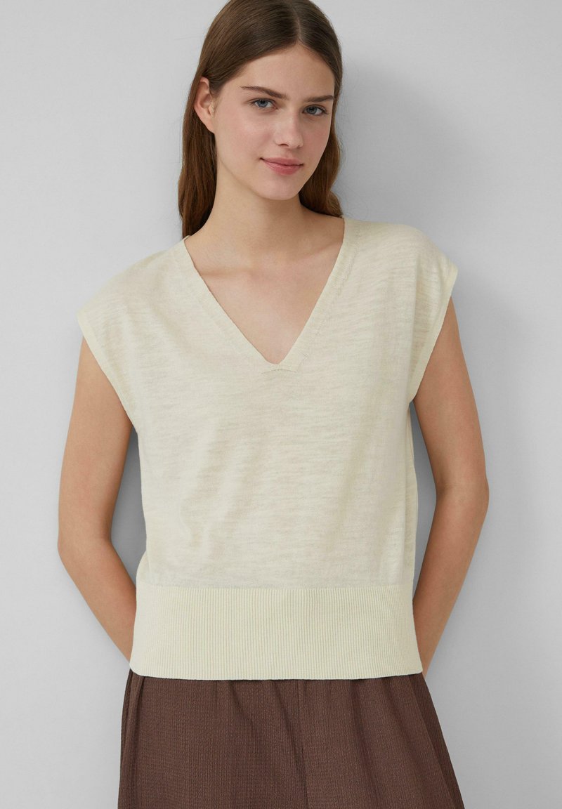 Pulower z dekoltem w serek i krótkim rękawem w jasnym kremowym kolorze, wykonany z teksturowanej tkaniny z prążkowanym wykończeniem. Połączony z brązowymi spodniami w kant.