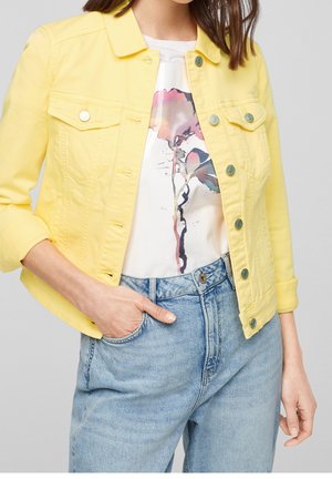 Woman wearing a light yellow buttoned jacket over a white floral t-shirt tucked into high-waisted light blue jeans with one hand in pocket.