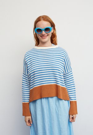 Lachende roodharige vrouw met felblauwe zonnebril, een blauw-wit gestreepte trui met bruine accenten en een lichtblauwe jurk.