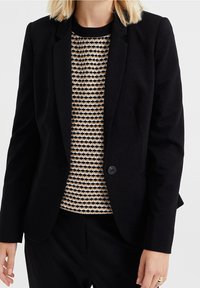 Svart skräddarsydd kavaj med enkel knappstängning och slagkrage, bärs över en mönstrad beige och svart tröja med ribbad halsringning.