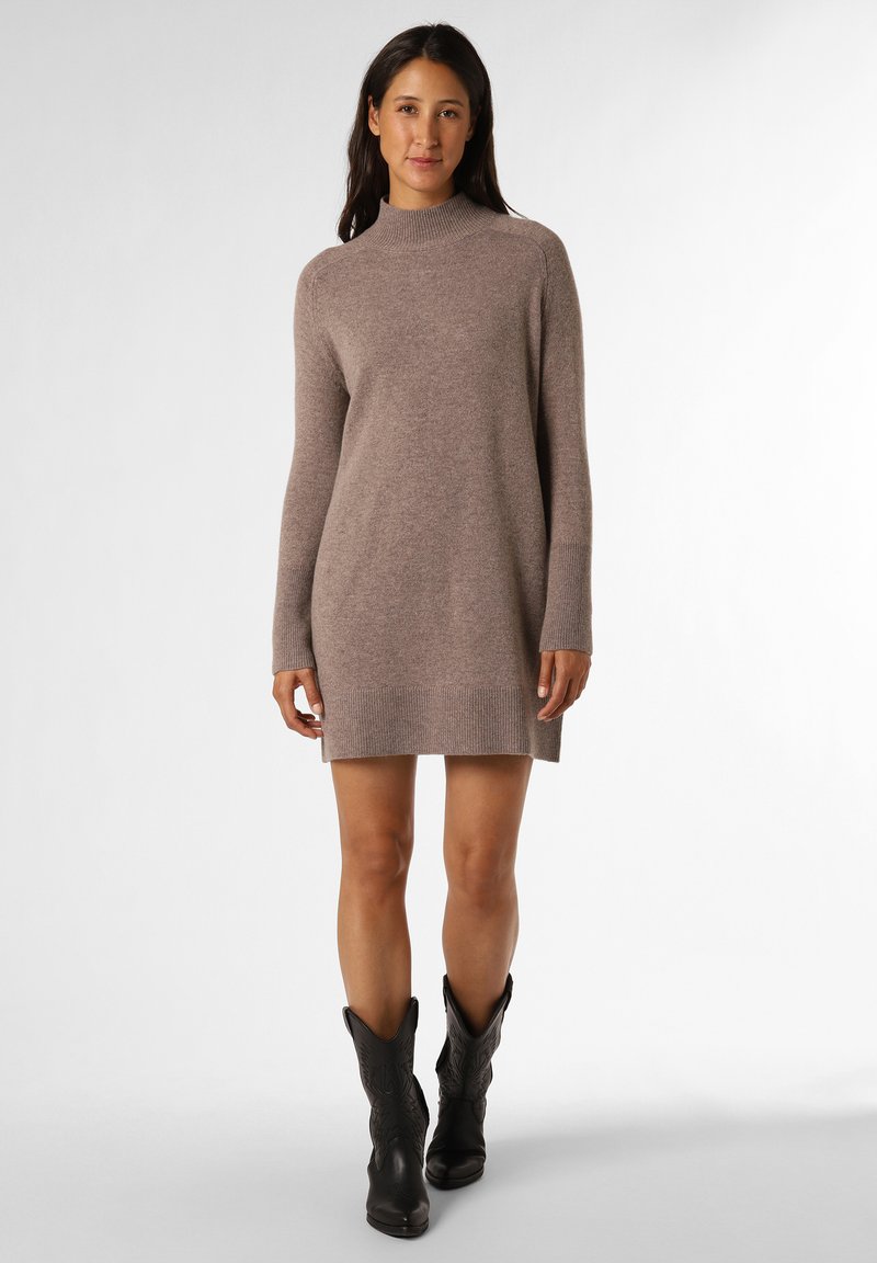 Strickkleid in hellem Taupe mit hohem Kragen, langen Ärmeln, gerippten Bündchen und weichem Material, kombiniert mit schwarzen Cowboystiefeln.