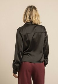 Chemise noire en satin à manches longues avec col et poignets boutonnés, présentant une texture lisse et un design élégant. Vue de dos.
