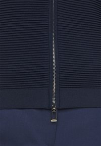 Granatowa bluza zapinana na zamek błyskawiczny z ribbowanym materiałem i gładką sekcją na dole. Posiada metalowy zamek z przodu.