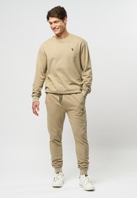 Man die glimlacht, gekleed in een beige sweatshirt en bijpassende joggers met witte sneakers, staande tegen een egale lichte achtergrond.