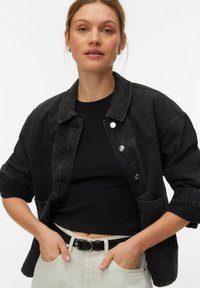 Crna traper jakna s ovratnikom, kopčanjem na gumbe i dva prednja džepa. Naočena preko crnog uskog topa, uparena sa svijetlim hlačama.