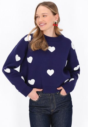 Maglione blu navale con motivi a cuore bianchi, colletto e bordo a coste, vestibilità rilassata, abbinato a jeans scuri. Texture in maglia morbida.