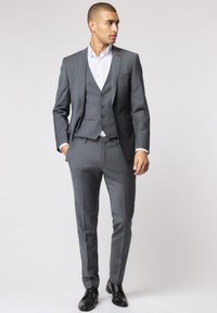 Grauer Dreiteiler mit schmaler Passform, strukturiertem Stoff, einreihiger Weste und maßgeschneiderter Hose. Kombiniert mit einem weißen Hemd und schwarzen Schuhen.