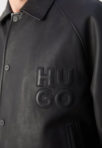 Melnas ādas jaka ar pogu aizdari, izteikta apkakle un iegravēta "HUGO" logo uz krūtīm. Gluda tekstūra un minimālisma dizains.