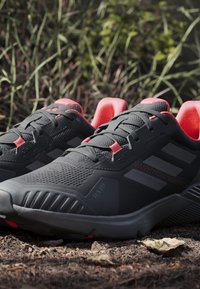 Scarpe da ginnastica nere con tomaia in mesh testurizzato, dettagli rossi e suole in gomma scura. Le caratteristiche includono lacci e un tallone imbottito.