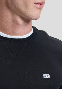 Close-up van een persoon die een zwart crewneck-sweatshirt draagt met een witte Lee-logo patch op de borst en een wit ondershirt.