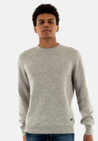 Jonge man met kort krullend haar in een eenvoudige lichtgrijze gebreide trui en een spijkerbroek, staande tegen een witte achtergrond.