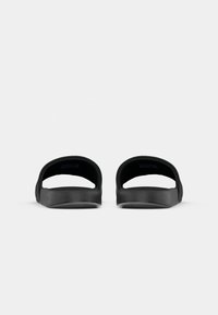 Paire de sandales à enfiler noires vues de face avec des semelles courbées et des larges brides supérieures sur fond blanc.