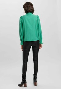Grüne Bluse mit gerafftem Ausschnitt, langen Puffärmel; kombiniert mit schwarzen, kunstledernen Hosen und schwarzen, hohen Stiefeletten.