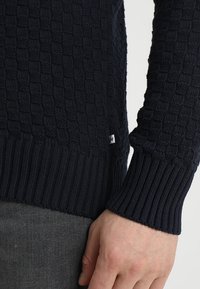 Maglione blu navy lavorato a maglia con un motivo a scacchiera testurizzato. Presenta polsini e orlo a coste, realizzato in tessuto morbido ed elasticizzato.