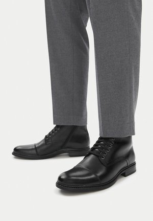 Czarne skórzane botki za kostkę z czubkiem w kształcie nakładki, gładką fakturą i pięcioma oczkami na sznurówki. Połączone z szarymi dopasowanymi spodniami.