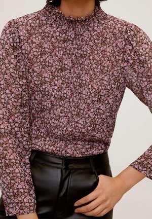 Femme portant un chemisier à manches longues rose et violet à motifs floraux avec un col montant froncé, rentré dans un pantalon en cuir noir taille haute.