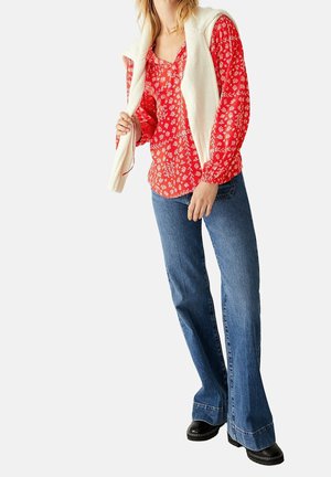 Blouse fleurie rouge à manches longues, associée à un jean bleu évasé et des bottines noires à la cheville ; pull en tricot crème drapé sur les épaules.