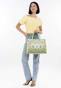 Borsa a mano con motivo geometrico verde e finitura lucida, caratterizzata da accenti neri a contrasto e doppi manici superiori, tenuta da una persona in abbigliamento casual.