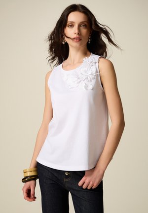 Donna con capelli scuri e ondulati indossa una canotta bianca con applicazioni floreali, jeans scuri, orecchini colorati e tre braccialetti impilati.