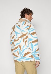 Giacca in pile morbido con un motivo di foglie nei colori beige, blu e bianco. Presenta un colletto alto e una vestibilità comoda, con tessuto testurizzato e cuciture visibili.
